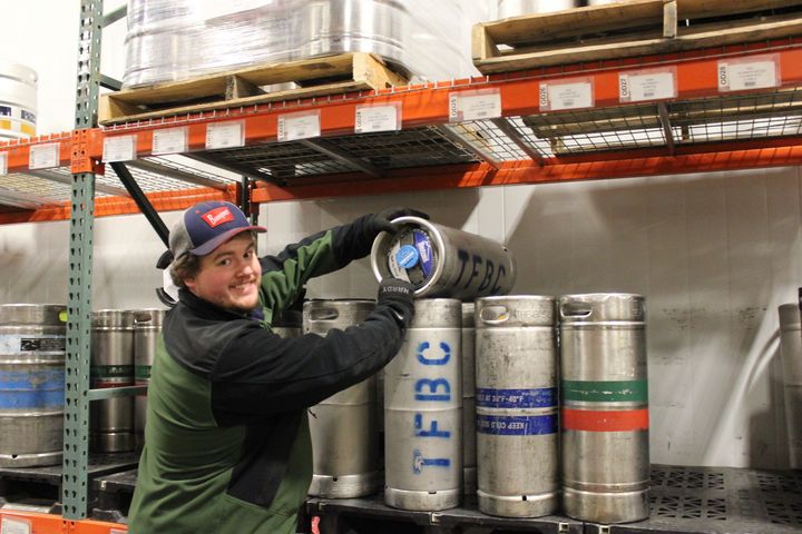 Worker stacking metal kegs on a warehouse shelf, wearing gloves and a cap, smiling at the camera