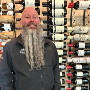 Smiling man with a long gray beard standing in front of a wall of wine bottles.