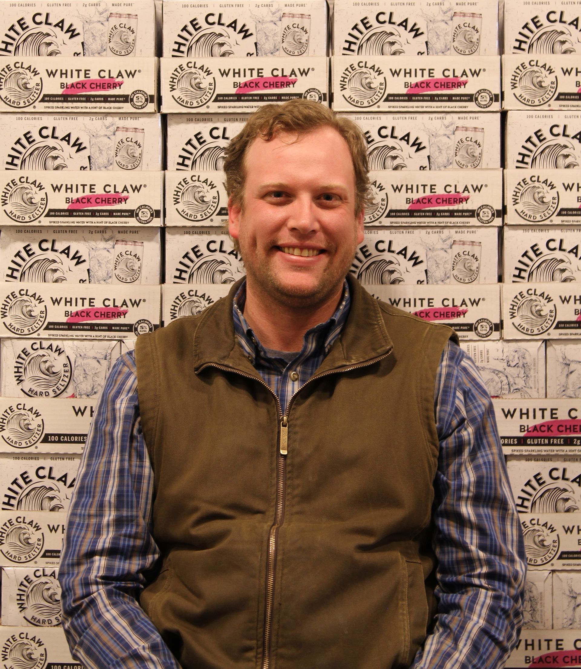 Smiling person in a blue plaid shirt and brown vest standing before a White Claw wall backdrop