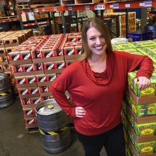Woman in red top smiling in a warehouse aisle beside stacked beverage displays and barrels.