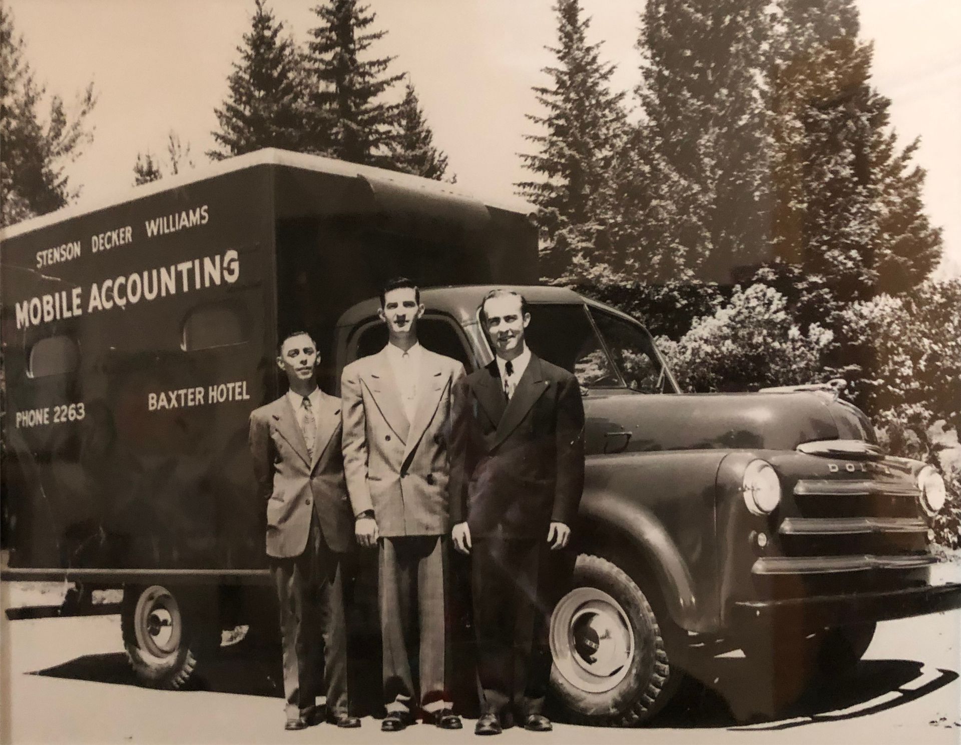 Three men stand beside a vintage mobile accounting truck with signs, parked outdoors by tall trees.