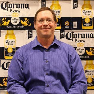 Man in a purple shirt smiling in front of a Corona Extra beer display.