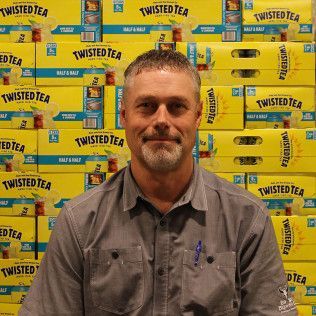 Man seated before a wall of Twisted Tea boxes, wearing a gray button-up shirt and faint smile
