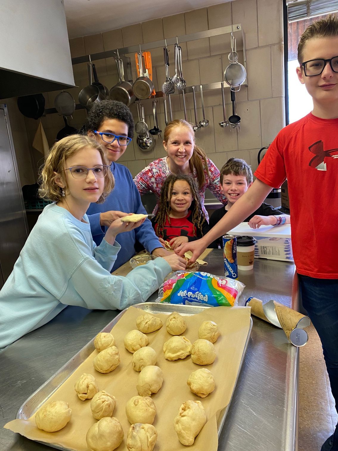 Children in a kitchen preparing and holding baked goods, smiling. Baking sheet on a counter.