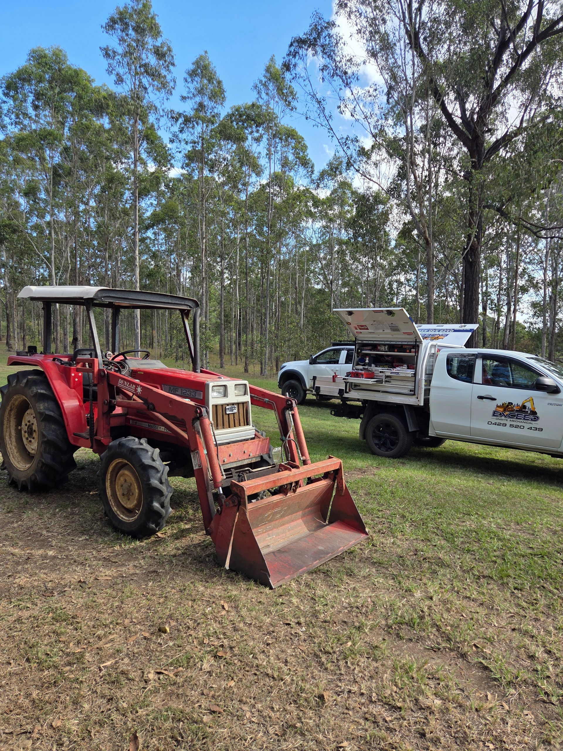 A red tractor with a front loader is parked on a grassy field in front of a white service truck — Exceed Onsite Mechanical in Grafton, NSW