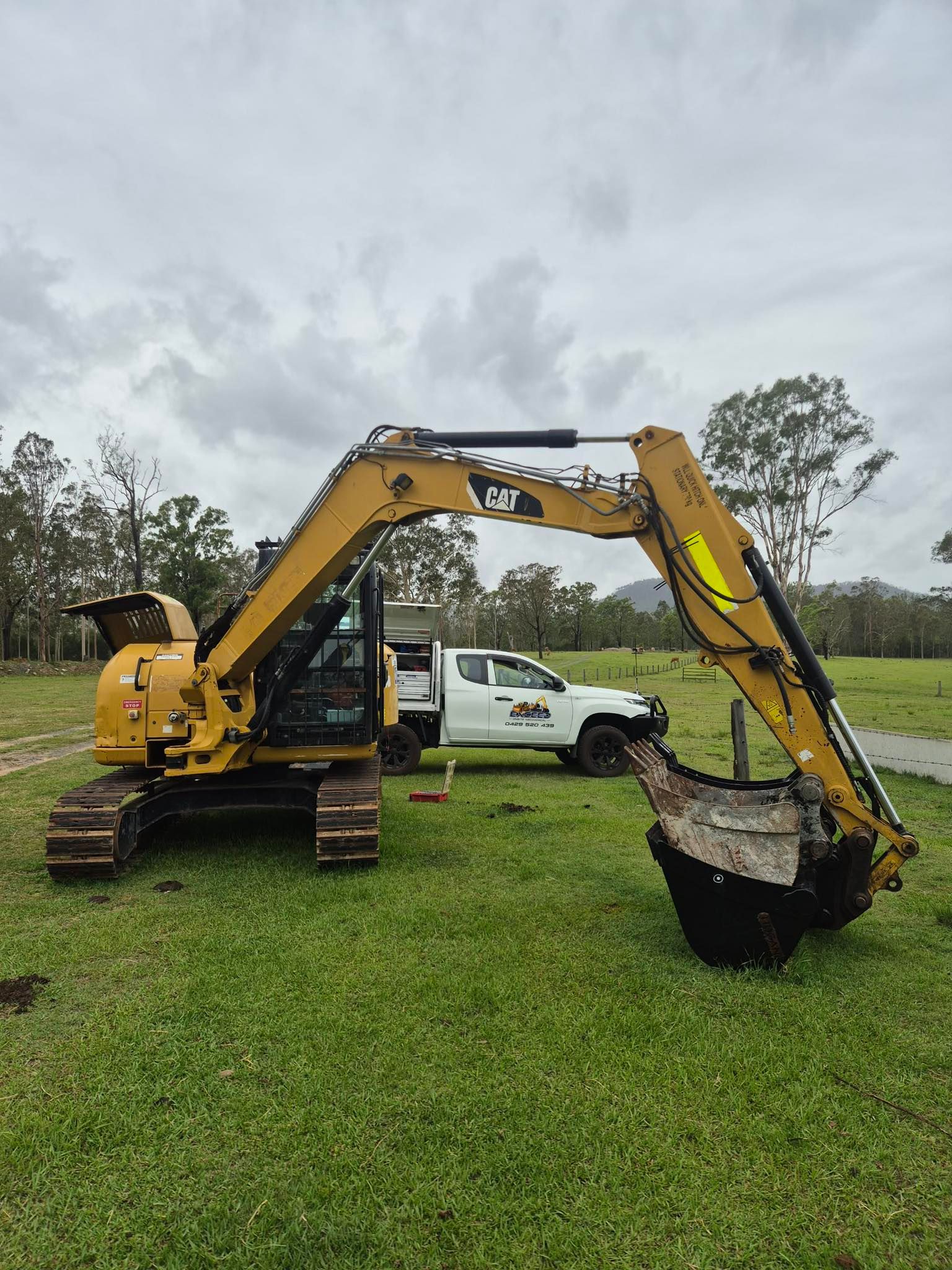 A Yellow Cat Excavator Sits in A Grassy Field with A White Pickup Truck Behind It — Exceed Onsite Mechanical in Grafton, NSW