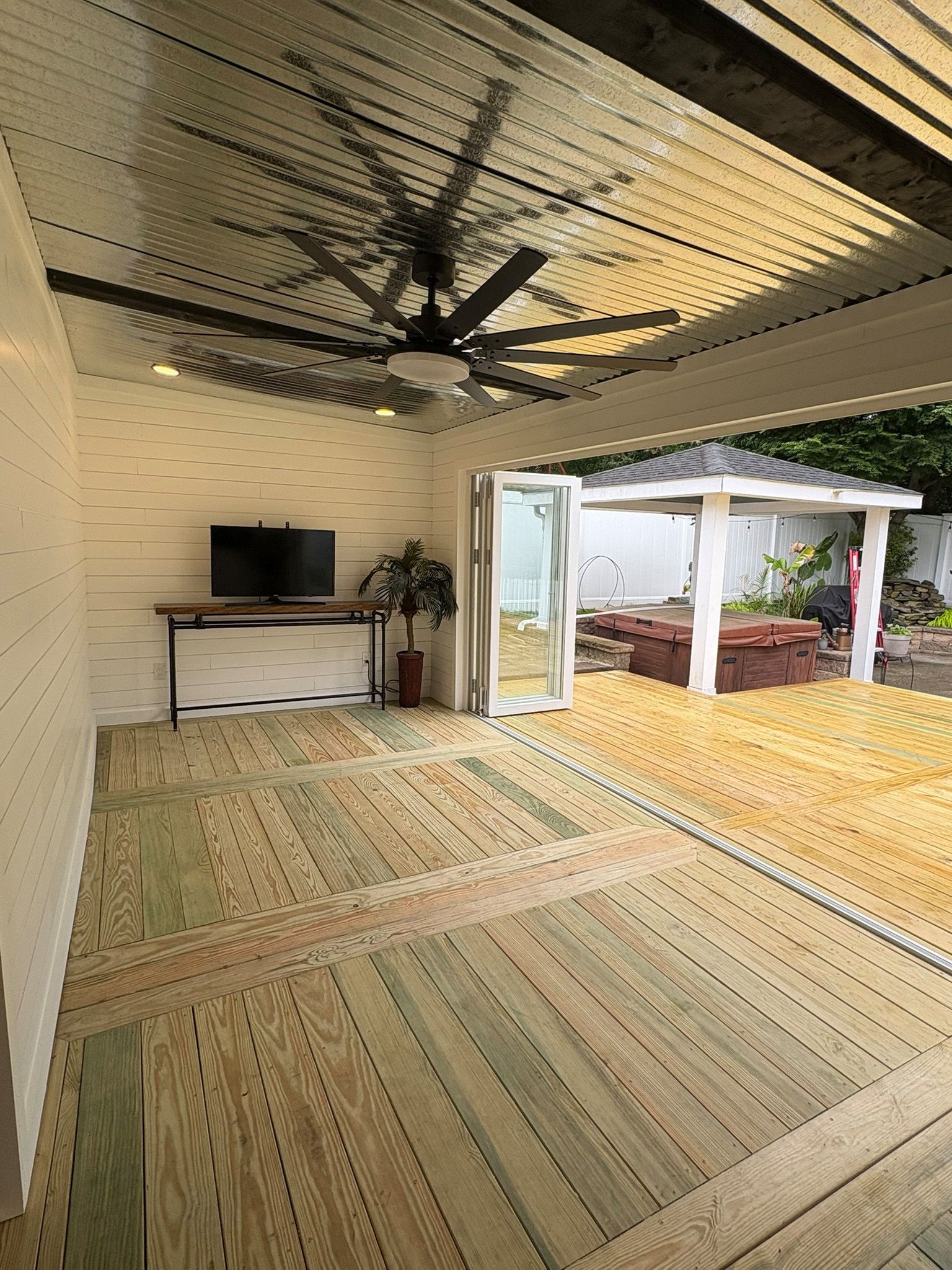 Outdoor patio with wood floor, ceiling fan, TV, and a view of a gazebo.