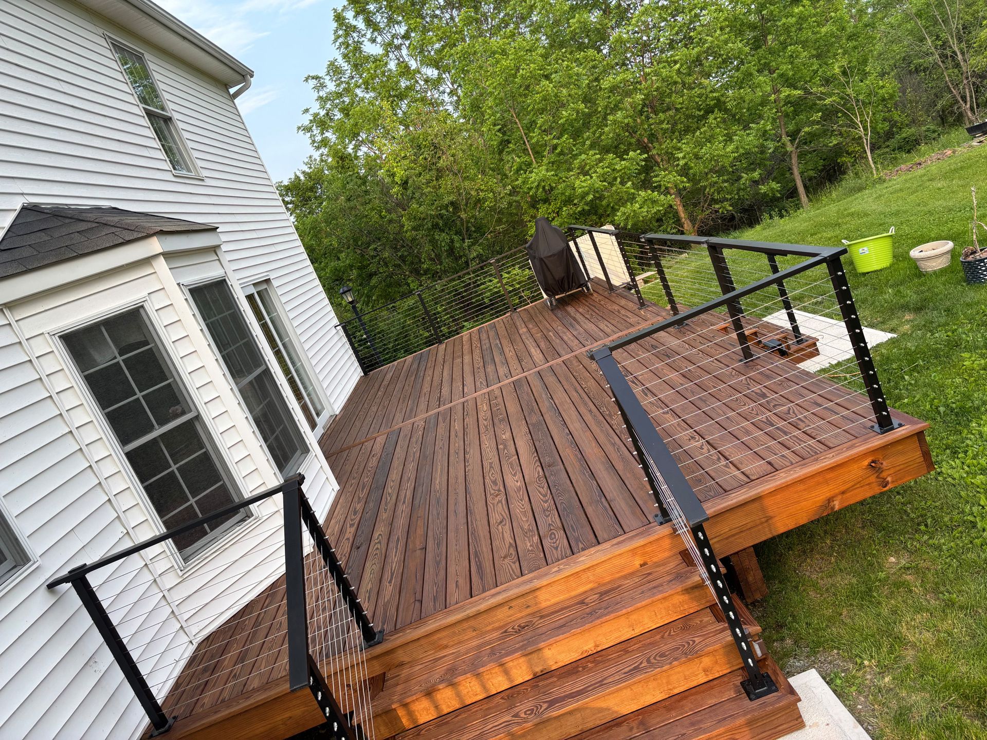Wooden deck with black railings, leading to steps. Green grass and a white house are visible.