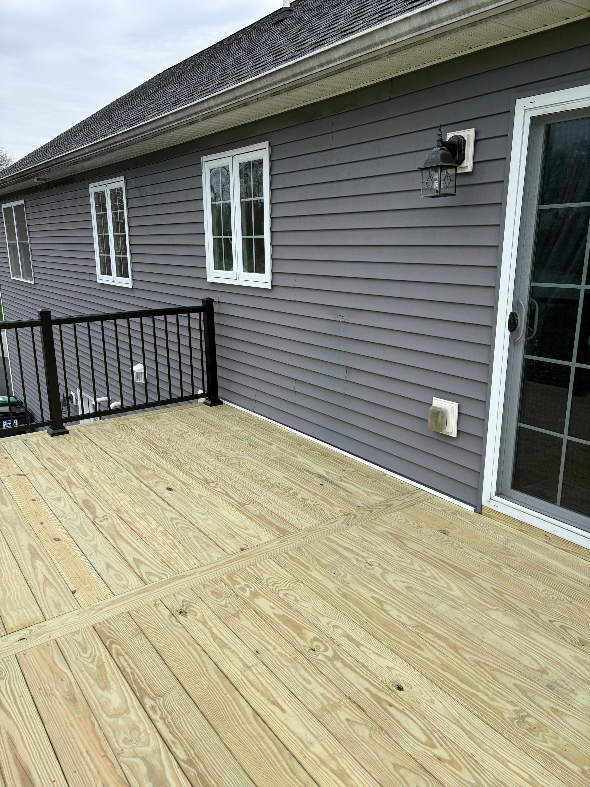 Exterior view of a deck with wooden planks, gray siding, black railing, and a sliding glass door.