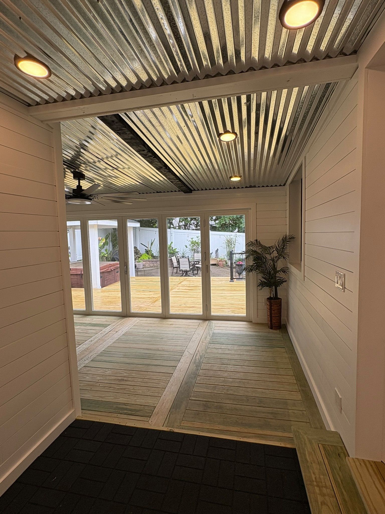 Hallway with corrugated metal ceiling, white walls, leading to a yard with folding glass doors and a patterned rug.