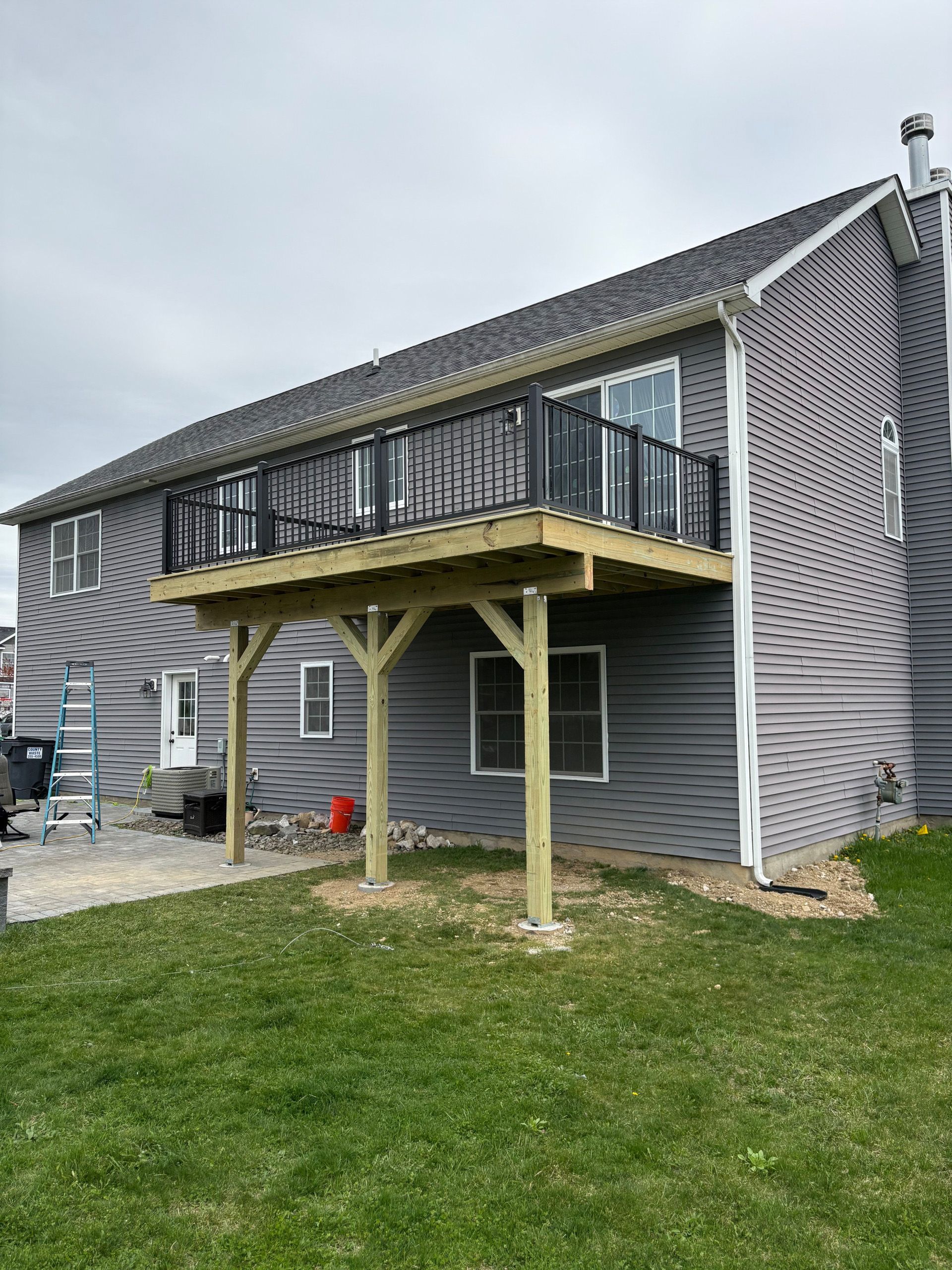 A two-story house with a wooden deck supported by posts. Black railing surrounds the deck.