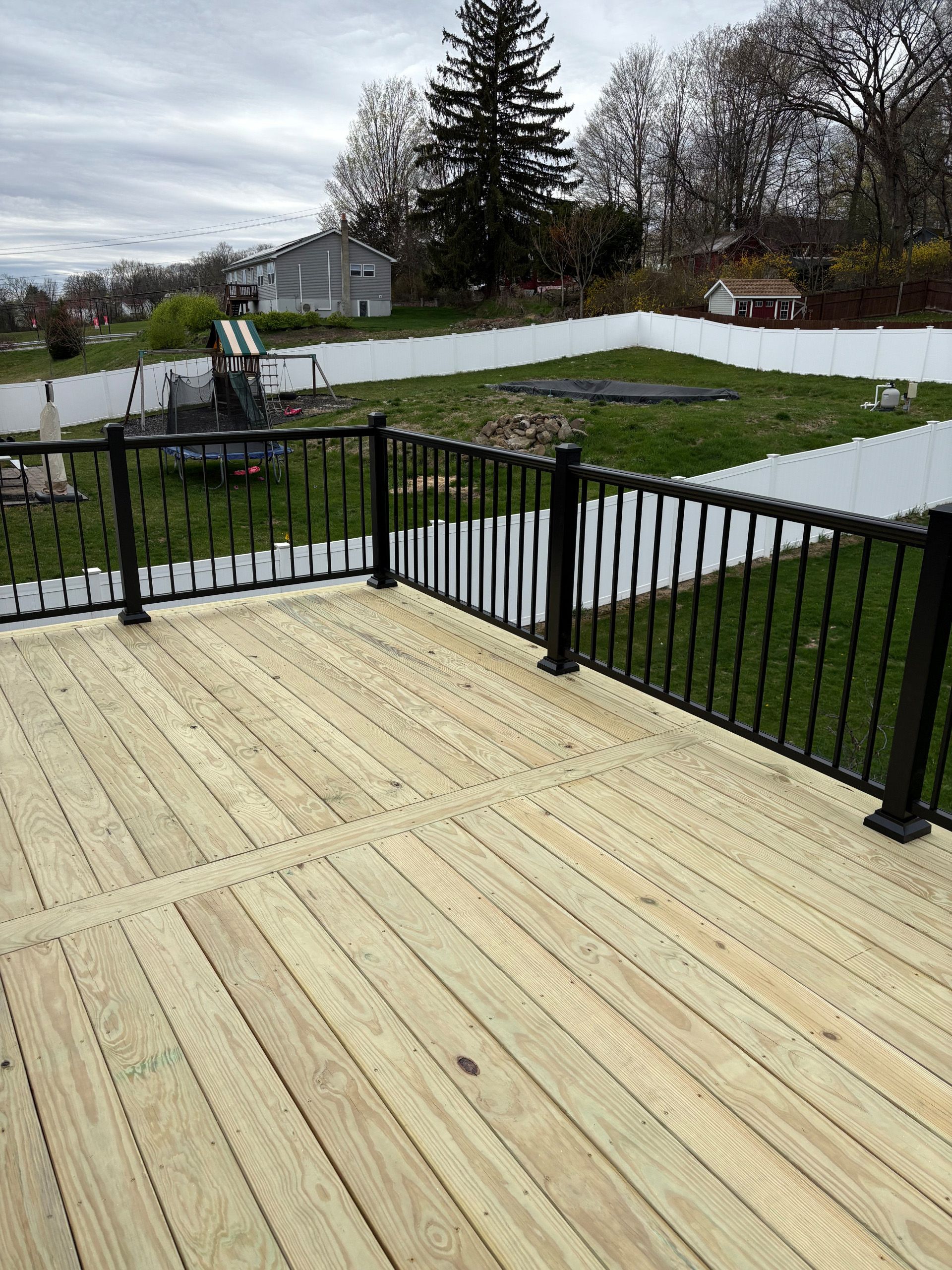 Wooden deck with black railing overlooking a fenced backyard, cloudy day.