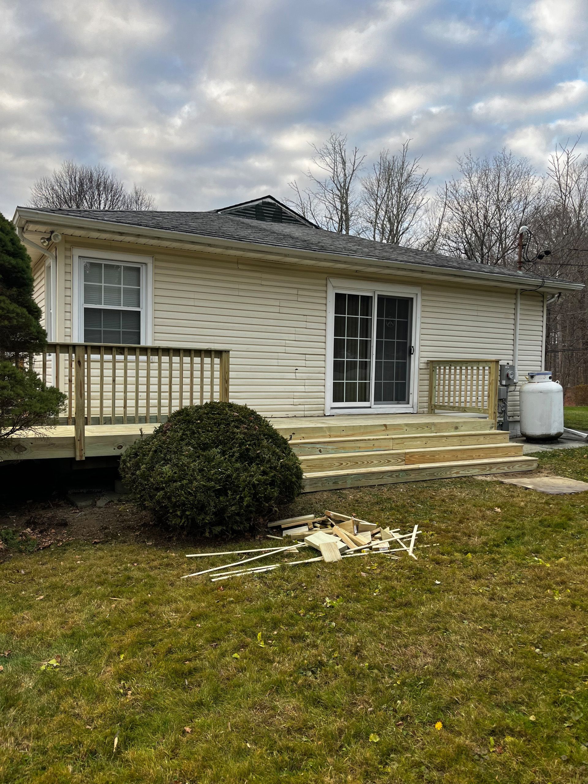 Small, beige house with a deck, sliding glass door, and propane tank. Cloudy sky, green grass.