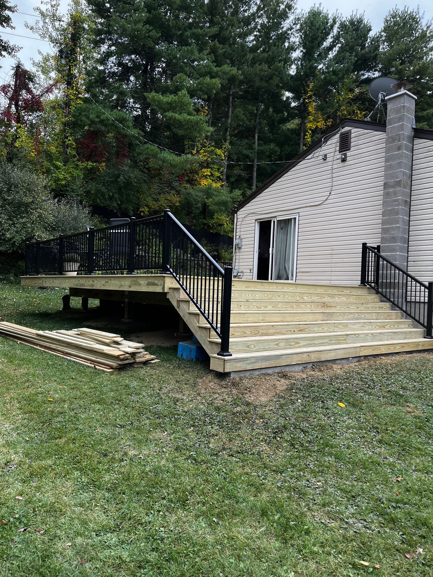 A wooden deck with black railings and steps leading to a house, surrounded by green grass and trees.