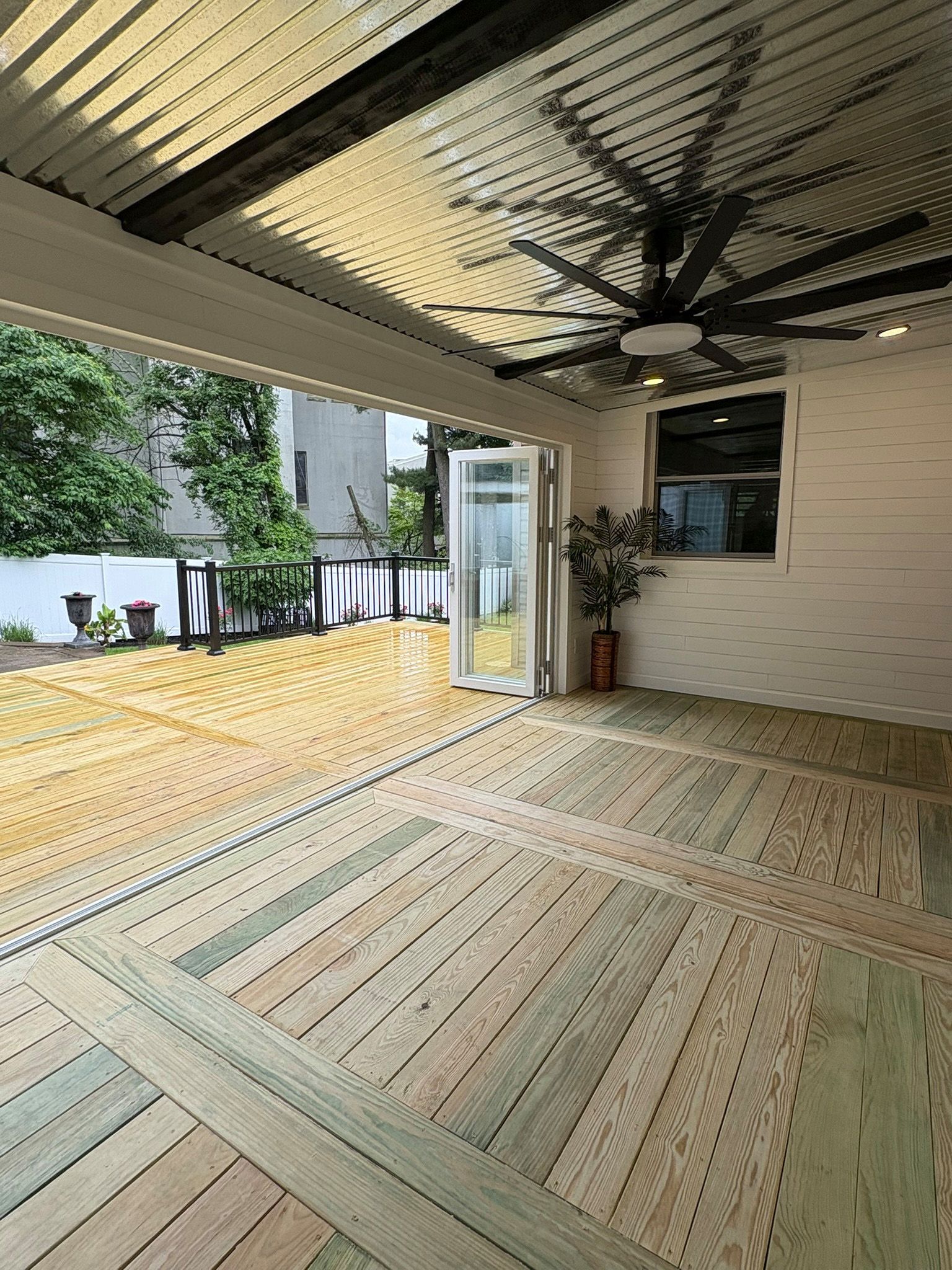 Covered outdoor deck with fan, wood flooring, folding glass doors, and a view of trees.