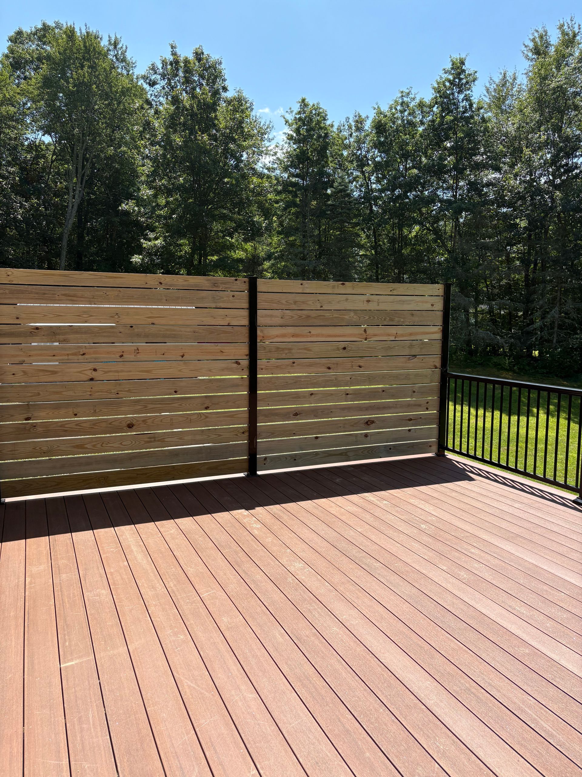 Wooden deck with a horizontal slat privacy fence, against a backdrop of green trees under a blue sky.