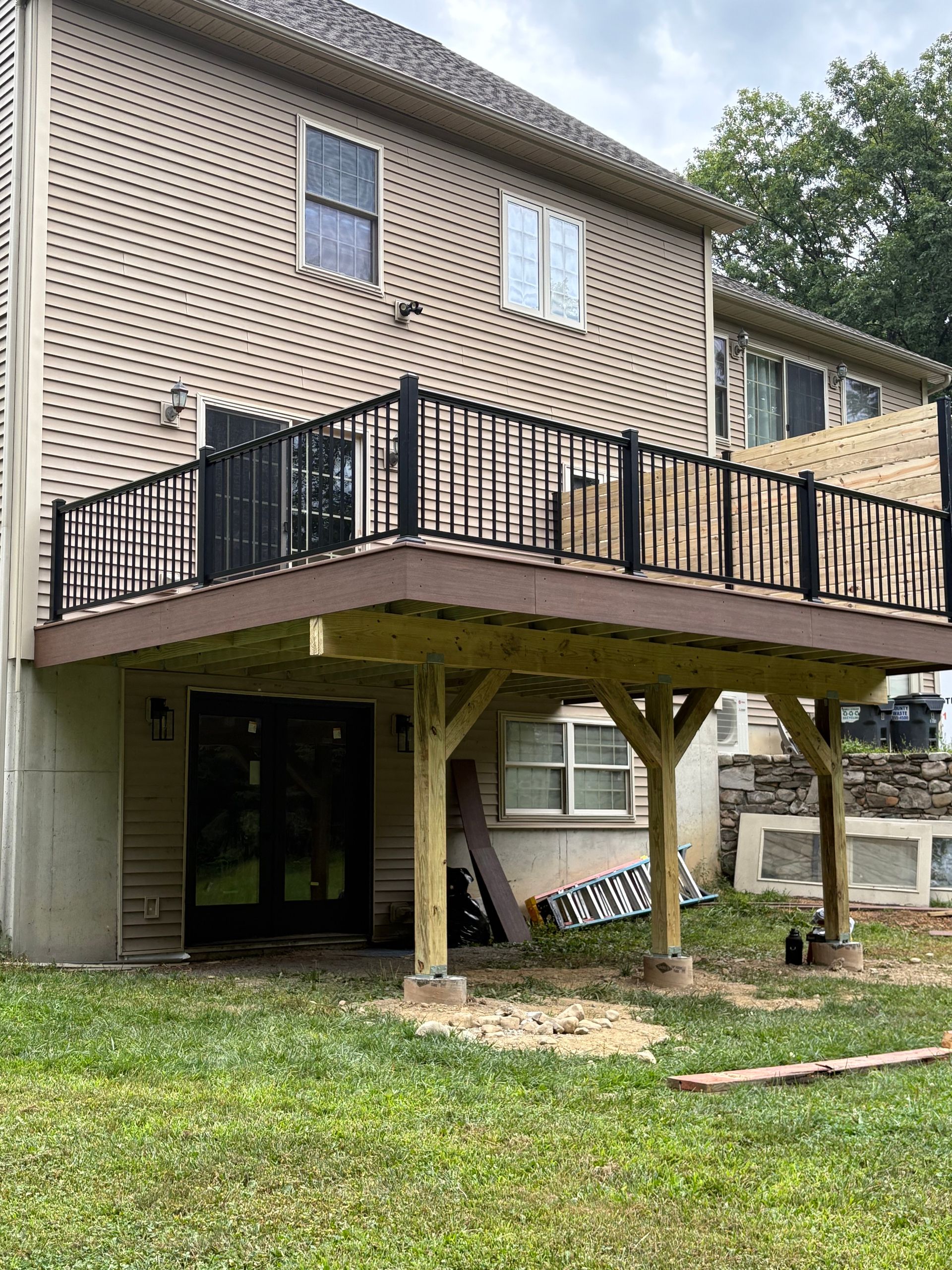 Two-story house with an elevated deck, black railing, and wooden supports on a grassy yard.