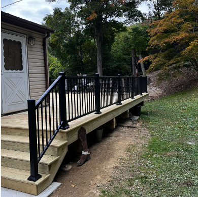 Wooden deck with black railings, steps leading up to a door, on a grassy hillside.