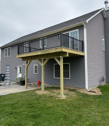 Elevated wooden deck with black railing attached to a gray house. Green grass and cloudy sky.