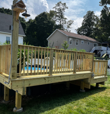 Wooden deck surrounding a pool, with a shower fixture and birdhouse on a post. Green grass and houses in the background.