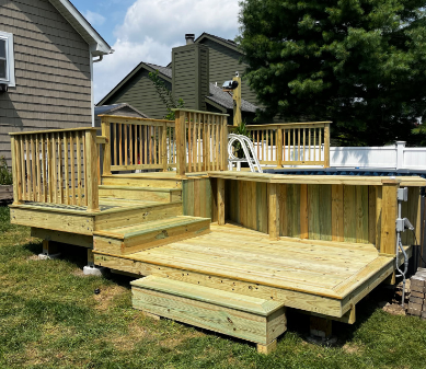 Wooden deck surrounds a pool with stairs, railing, and a storage bench in a backyard.