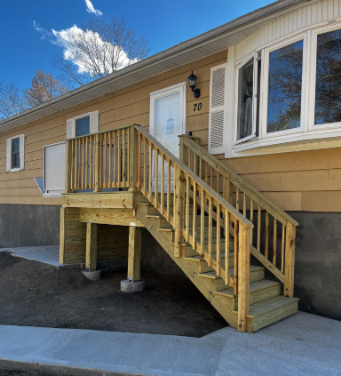 Wooden stairs leading up to a light yellow house with a white door and a bay window.