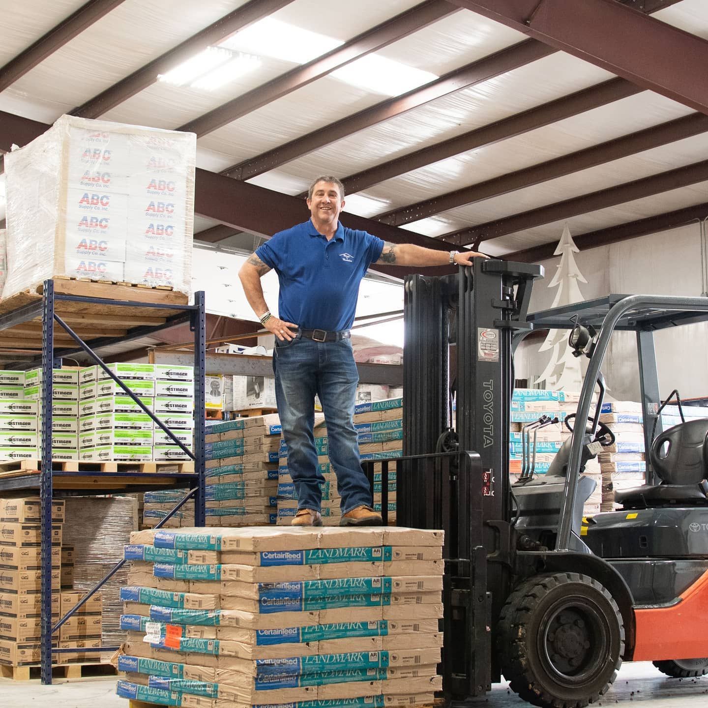 Dan Nicholas, owner of Roofs by Nicholas, standing on roofing material stacks in warehouse with forklift