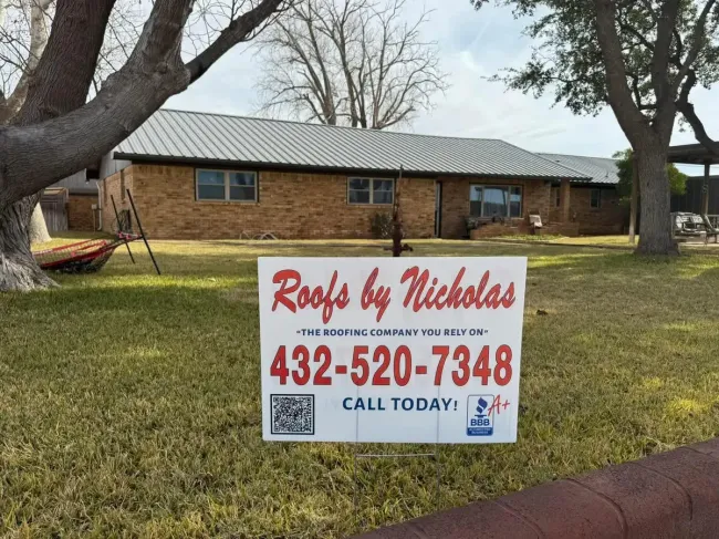 Roofing yard sign on a lawn in front of a brick house with a new metal roof, reading “Roofs by Nicholas” and phone number 432-520-7348.