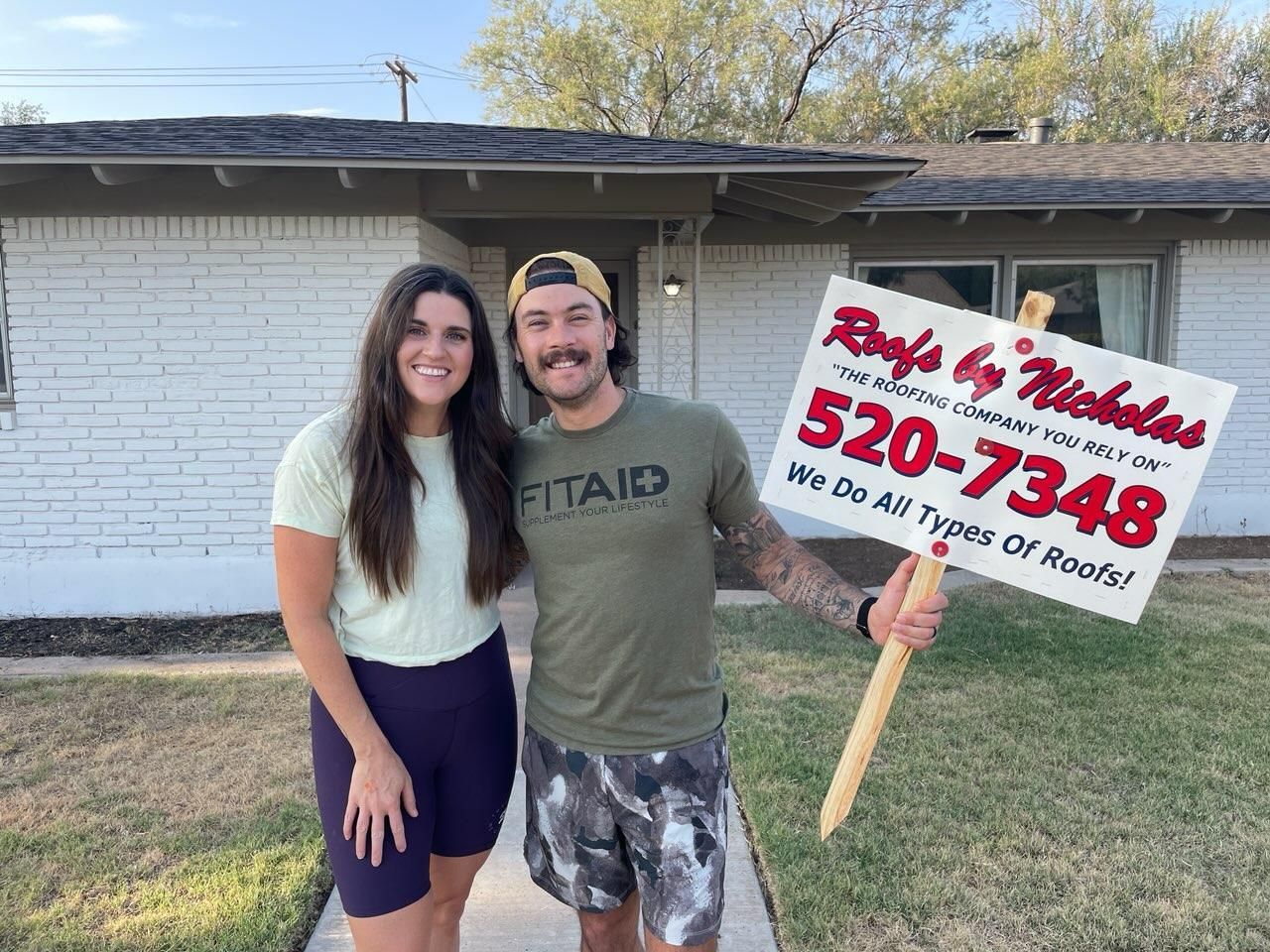Happy couple standing outside their home holding Roofs by Nicholas sign after roof service