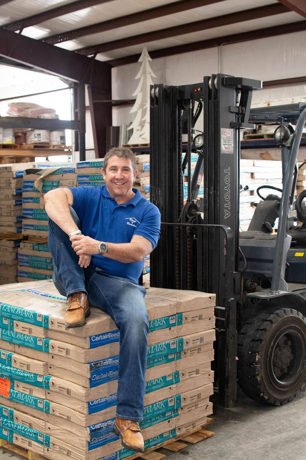 Dan Nicholas, owner of Roofs by Nicholas, sitting on roofing material stacks in warehouse with forklift in background