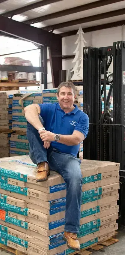 Dan Nicholas, founder, sitting on stacked pallets beside a forklift in a warehouse, wearing a blue shirt and jeans.