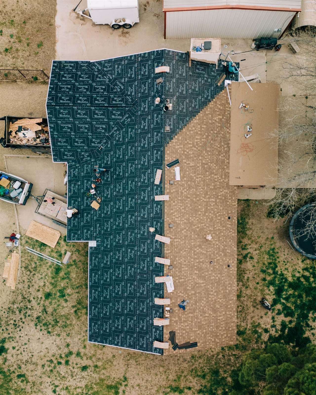 Overhead drone view of a Roofs by Nicholas crew installing tan shingles over branded synthetic underlayment, with tear-off debris in a trailer below