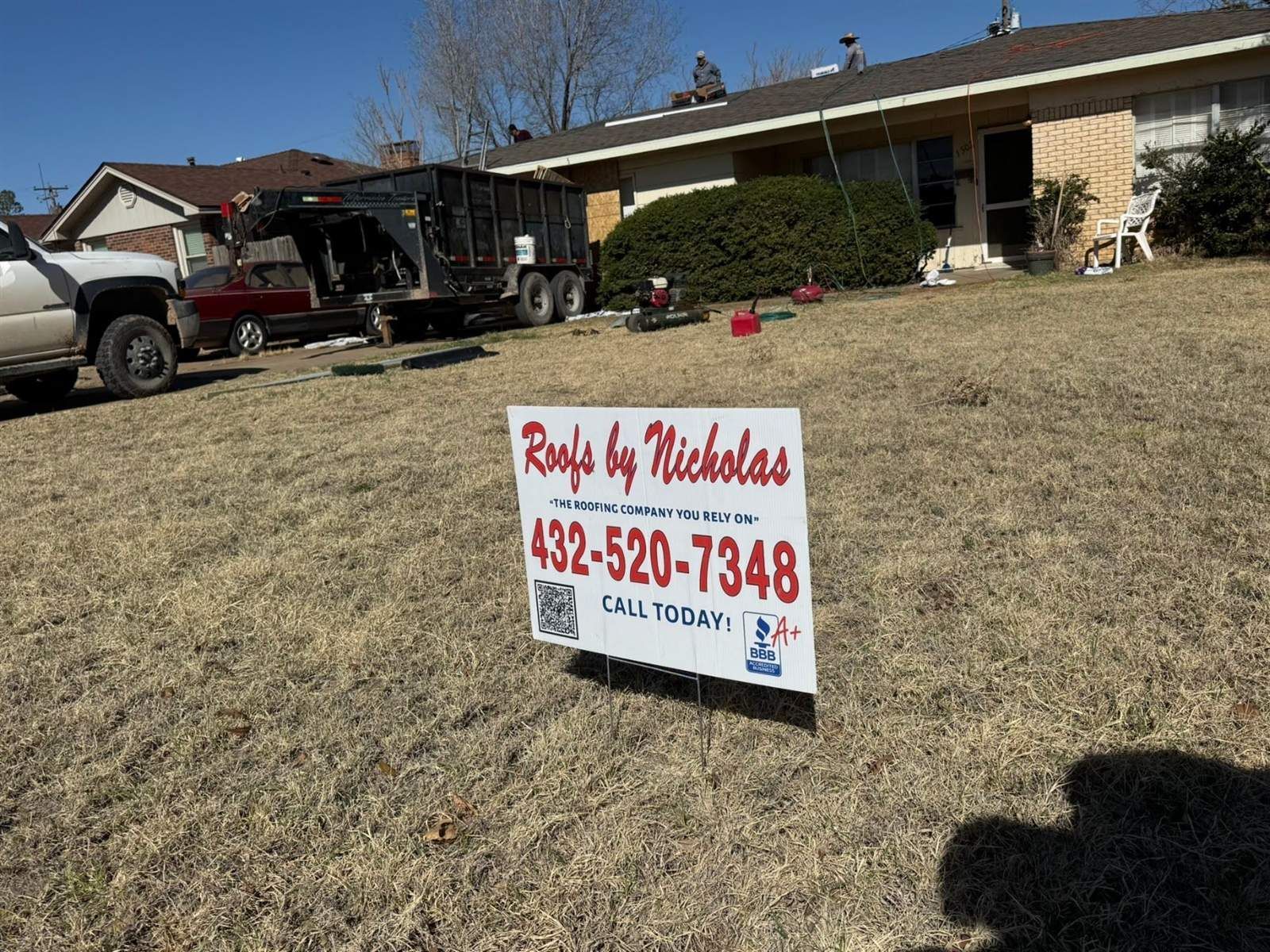 Roofs by Nicholas yard sign at a brick home during a roof replacement, with crew on the roof and a dump trailer in the driveway
