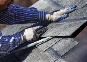 Roofer cutting grey asphalt shingles during roof installation with utility knife