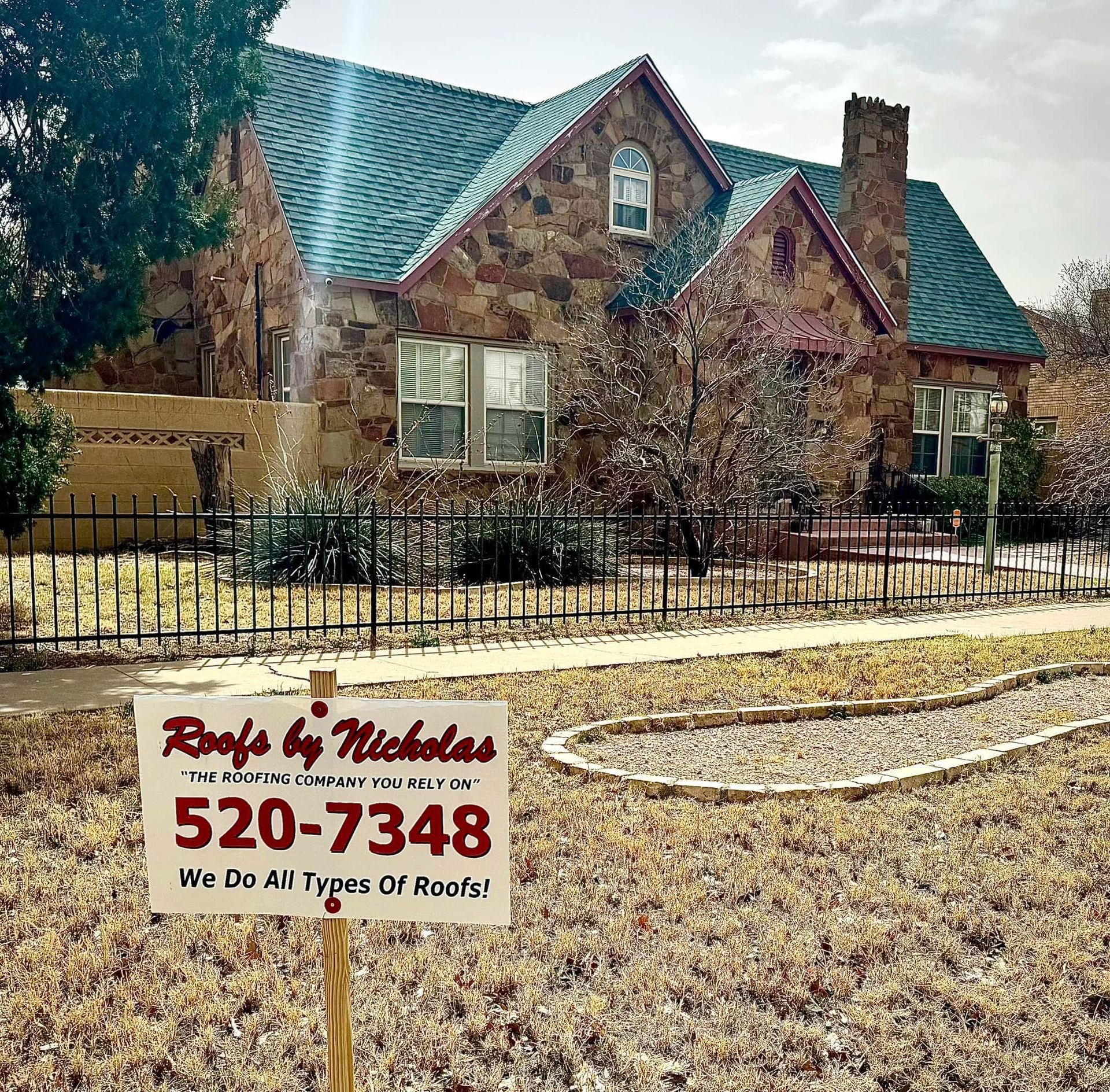 Roofs by Nicholas sign in front of a stone house with a steep green roof in Midland Texas