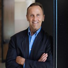 A man in a suit and blue shirt is leaning against a wall with his arms crossed.