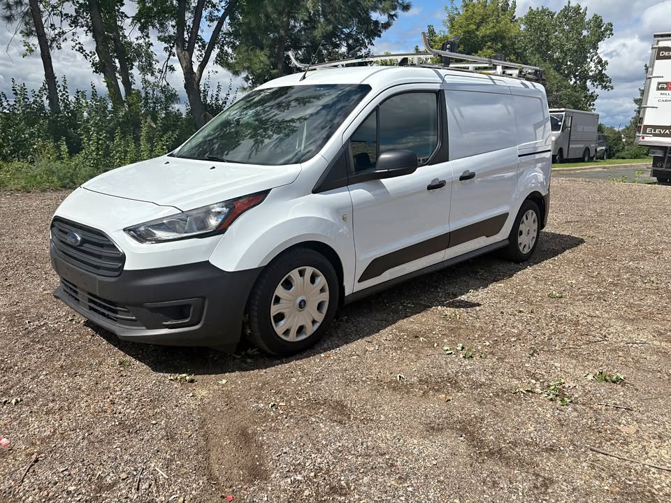 White Ford Transit Connect van parked on gravel, with ladder rack on the roof.