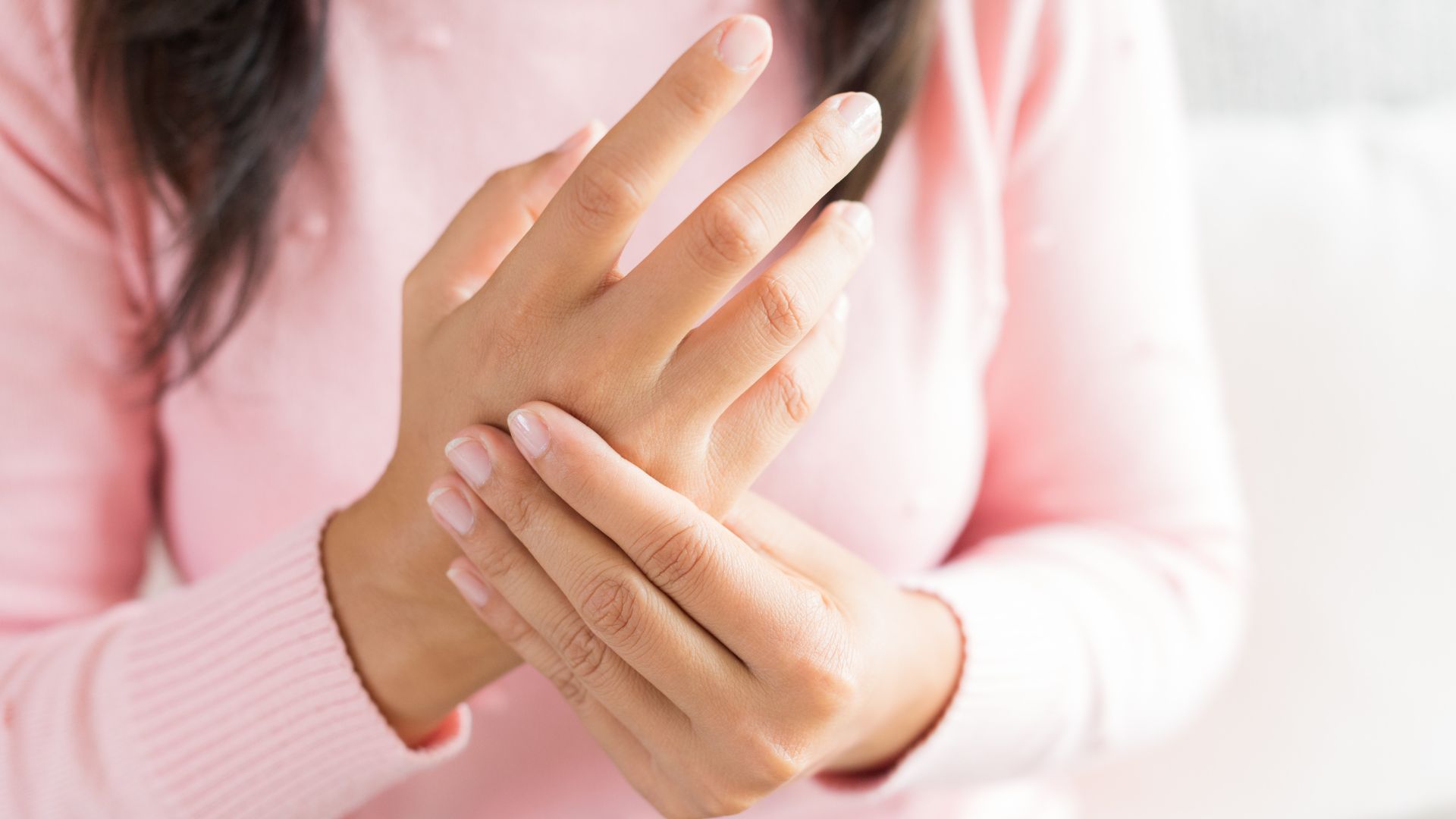 Woman in pink sweater holding hands, examining fingers, likely for pain or swelling.