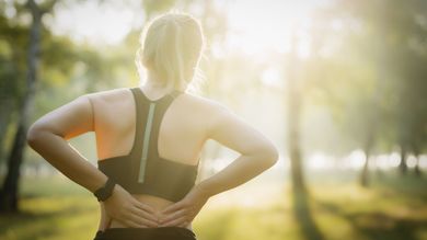 Woman in workout clothes holds her lower back outdoors, possibly in pain, with sunlight.