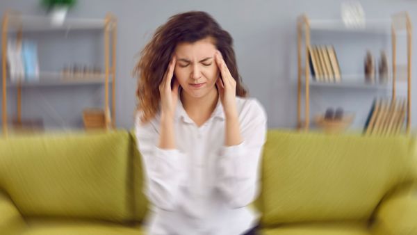 Woman with hands on temples, eyes closed, appearing to have a headache, blurred background of a living room.
