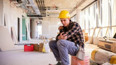 Construction worker in yellow hard hat, holding arm, sitting on bucket in unfinished building.