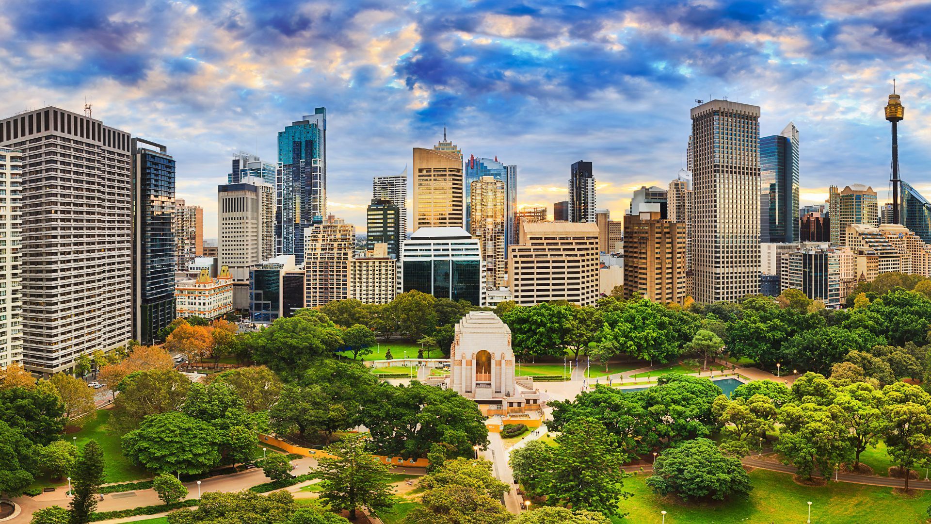 City skyline with park in foreground under a cloudy sky. Buildings of various heights and materials.