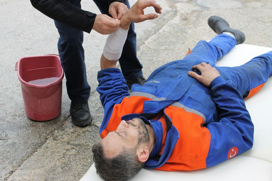 Person wrapping bandage on injured arm of another person lying down outside. A bucket of water sits nearby.