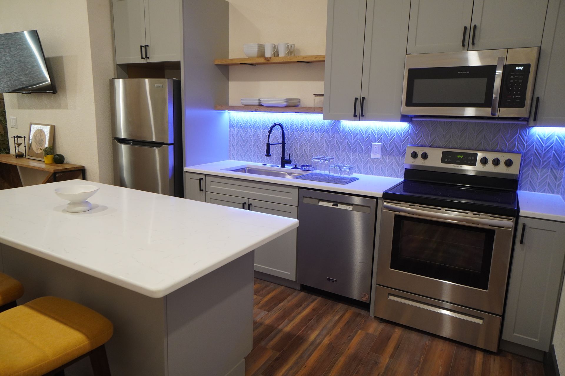 Modern kitchen with stainless steel appliances, gray cabinets, white countertop, and blue under-cabinet lighting.
