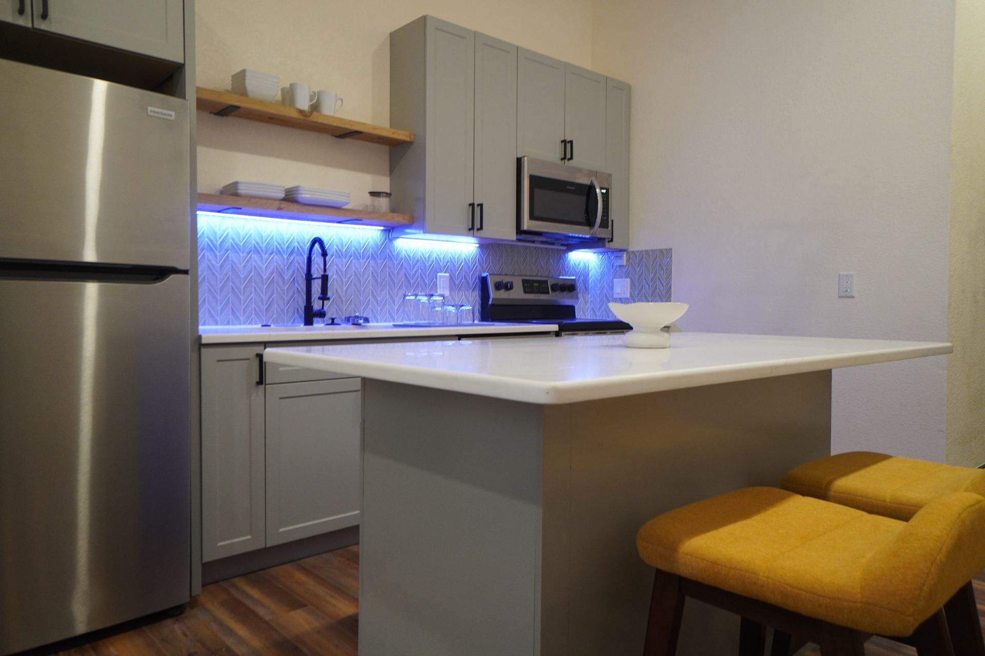 Modern kitchen with grey cabinets, stainless steel appliances, island with yellow stool, and blue under-cabinet lighting.