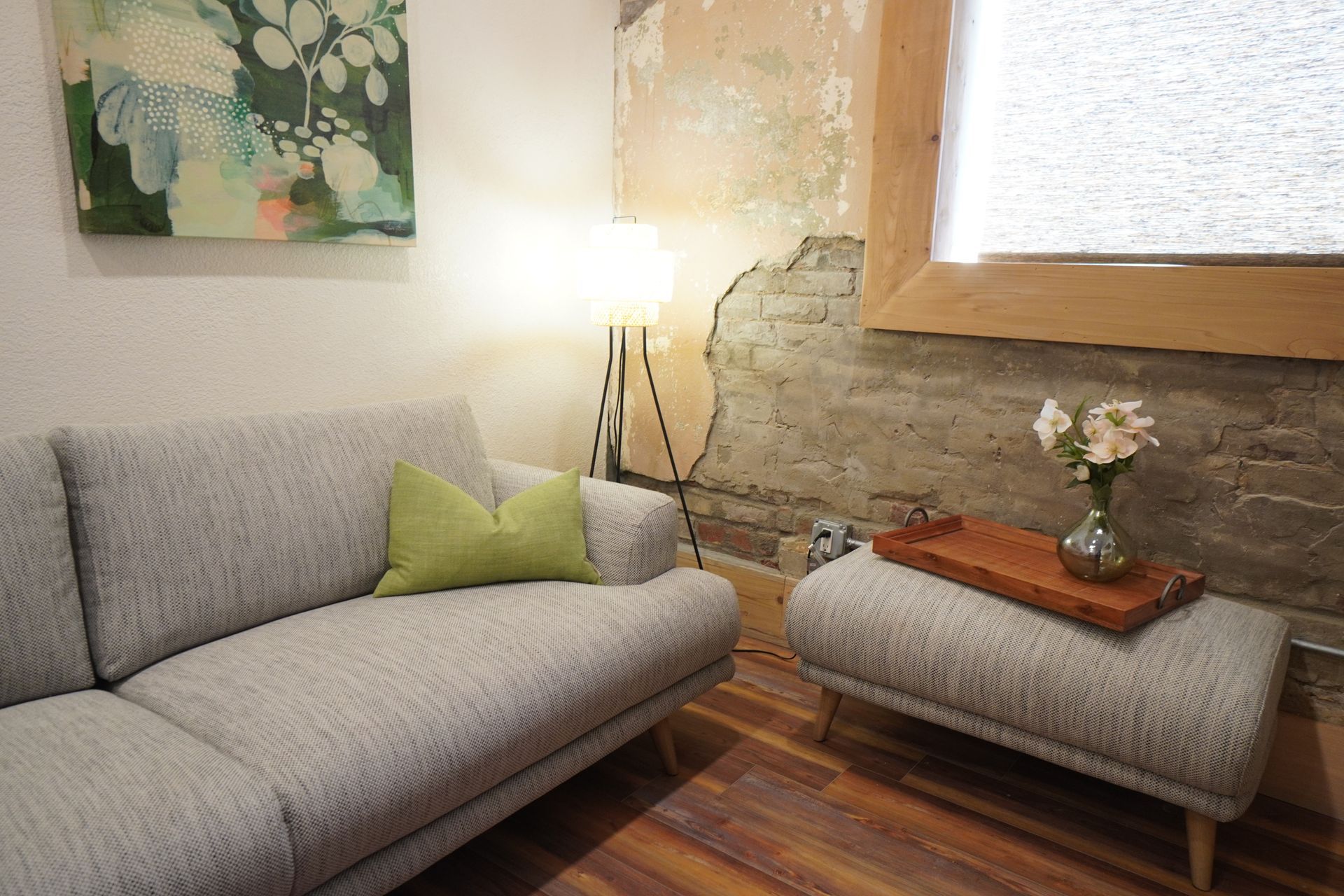 Gray sofa with green pillow, ottoman, artwork, and lamp in a room with a brick wall and flowers.