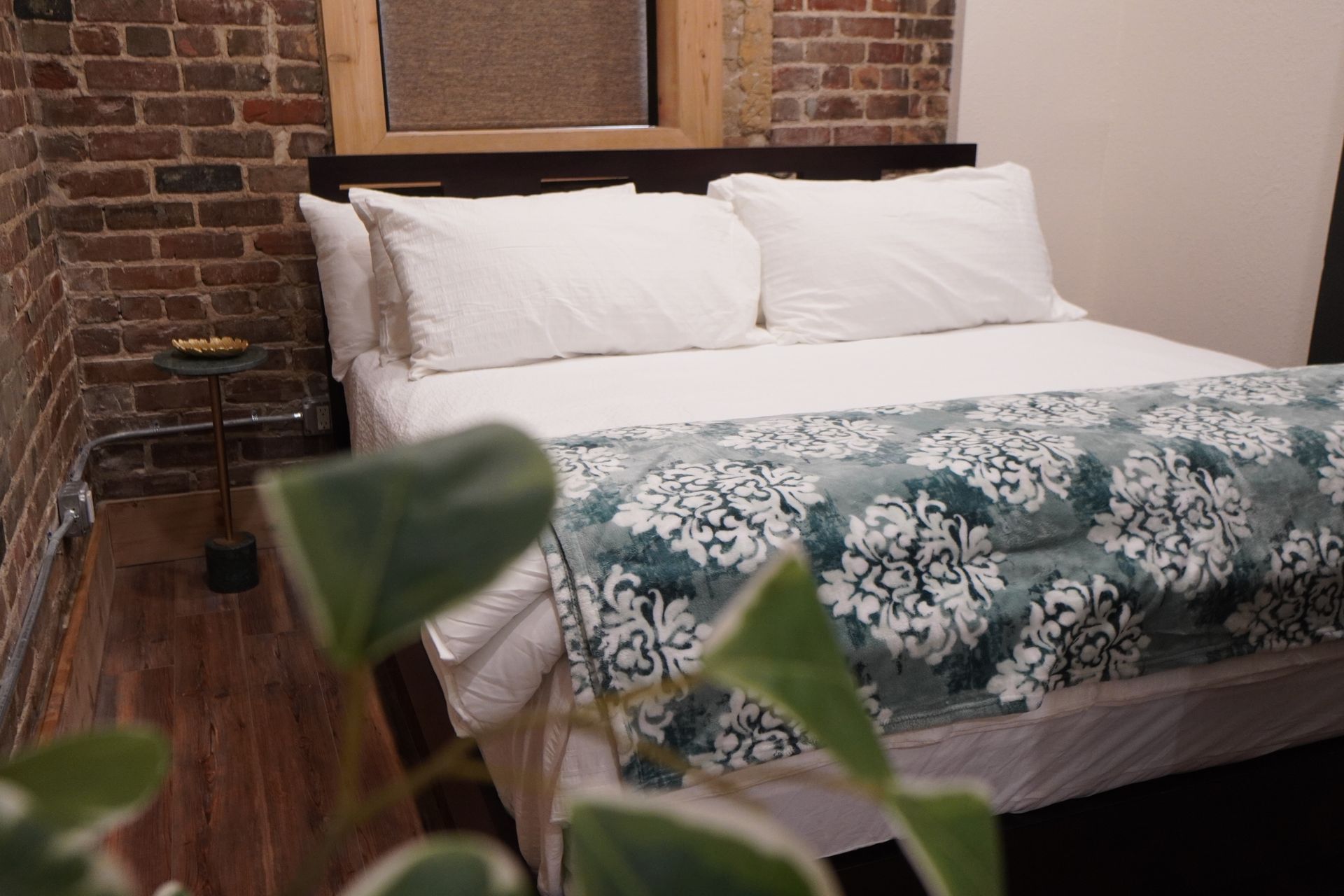 Bedroom with white bedding, patterned blue blanket, brick wall, small side table.