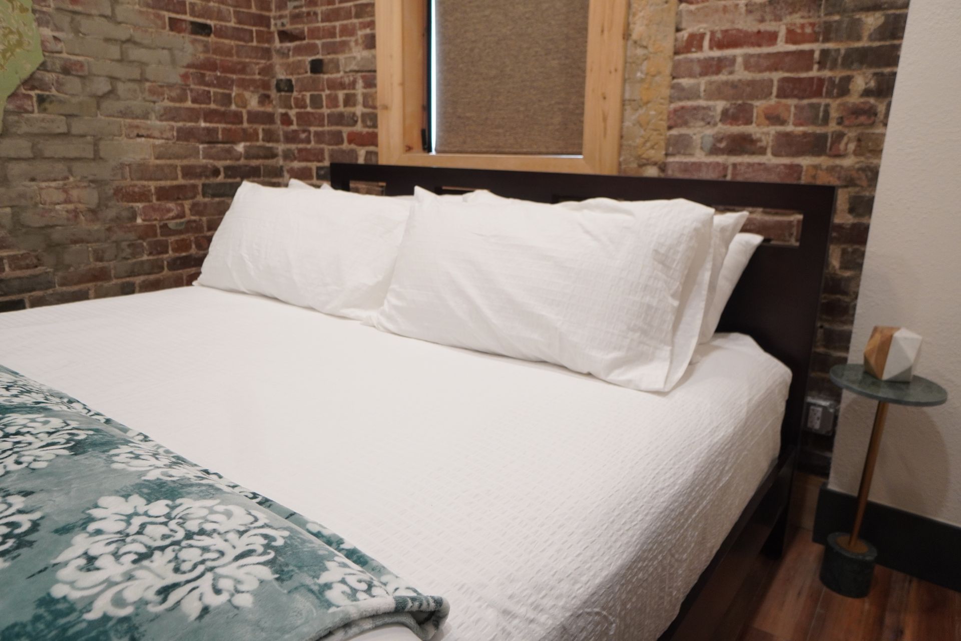 Bedroom with a made bed, white linens, brick wall, window, and small side table.