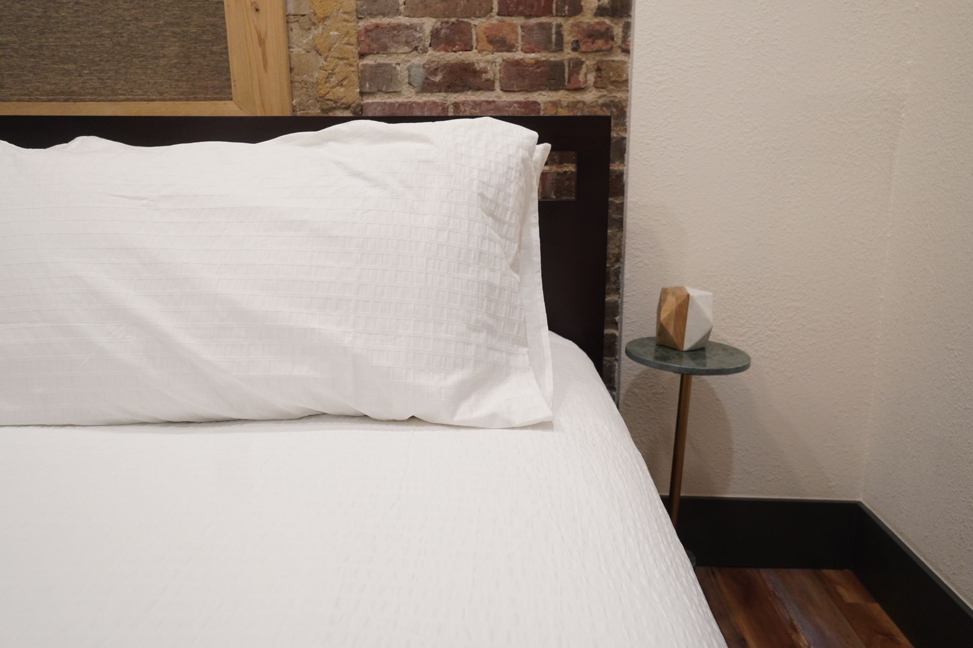 White bed with a pillow, small table, and a decorative object. Brick wall and wood floor.