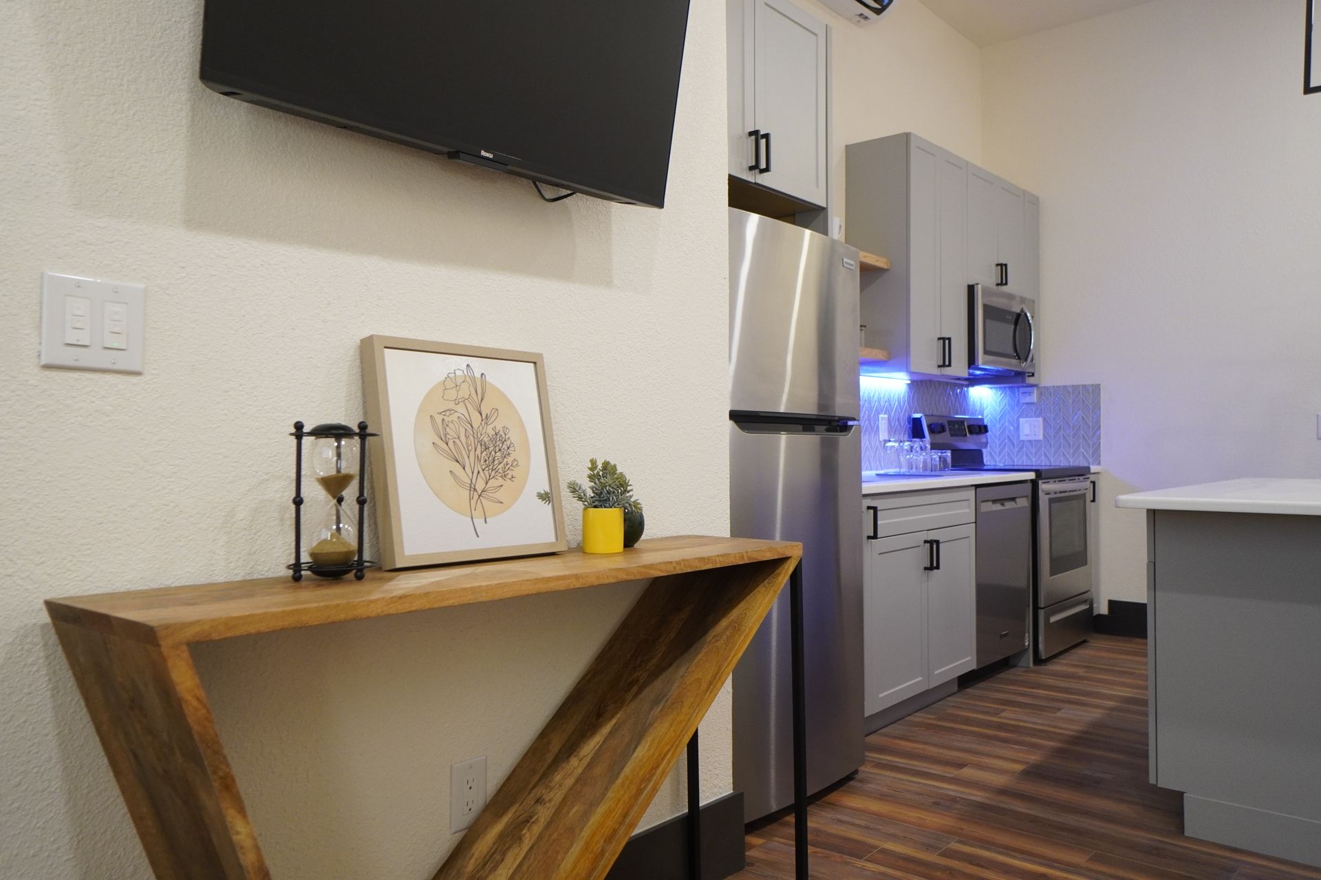 Wooden console table with decor against a wall, leading into a gray kitchen with stainless steel appliances.